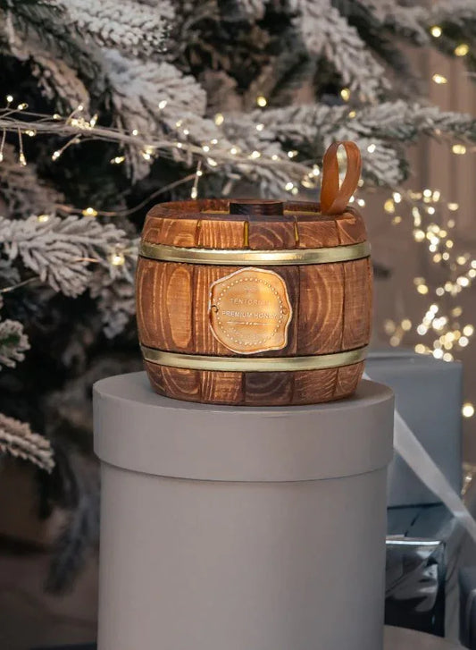 Wooden barrel-shaped honey jar with a gold label on a gray pedestal, Christmas tree in the background