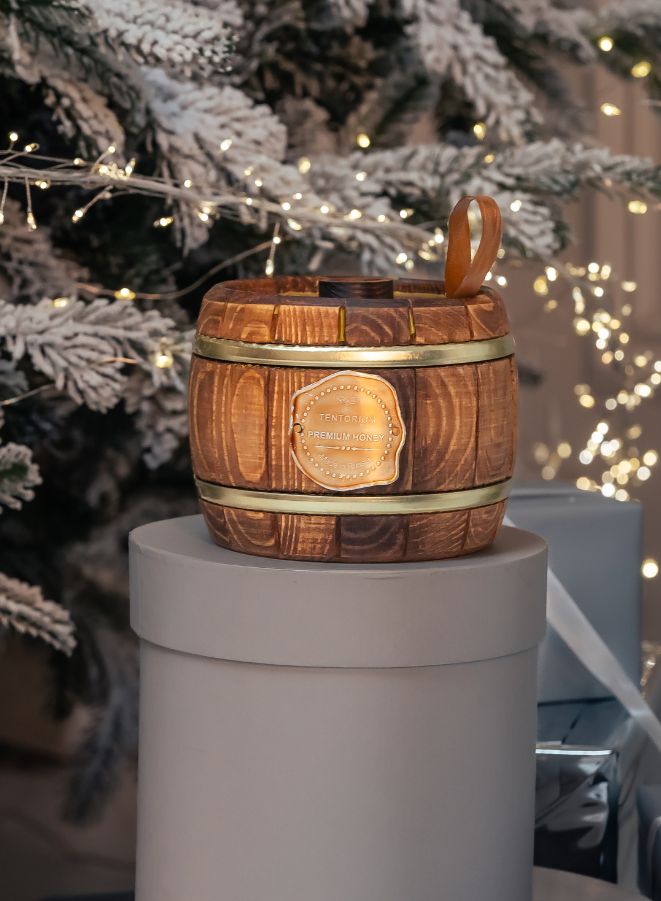 Wooden barrel-shaped honey jar with a gold label on a gray pedestal, Christmas tree in the background