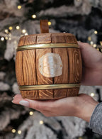 Wooden barrel-shaped ornament with gold band held in a hand against a blurred Christmas tree background.